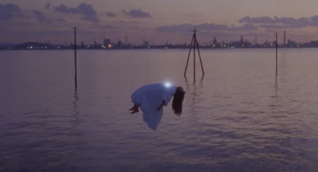 Ichiko Aoba screen shot from the music video for Sonar. The image shows a woman with a glowing heart levitating above a body of water at dusk.