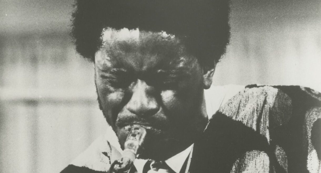 Black-and-white photo of Pharoah Sanders in 1968, eyes clenched shut in deep concentration as he plays tenor saxophone. His face is glistening with sweat, and he wears a boldly patterned garment over a collared shirt and tie. The image captures an intense, emotional moment mid-performance, highlighting the physicality and spiritual commitment of his playing.