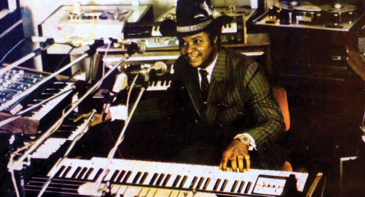 A man, William Onyeabor, in a pinstripe suit and cowboy hat smiles while seated at a large keyboard surrounded by synthesizers, microphones, and recording equipment in a studio.