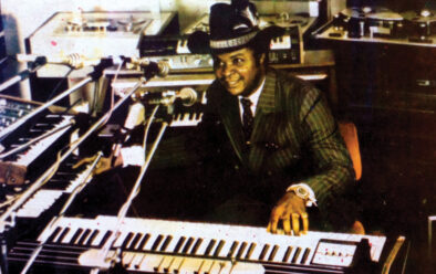A man, William Onyeabor, in a pinstripe suit and cowboy hat smiles while seated at a large keyboard surrounded by synthesizers, microphones, and recording equipment in a studio.
