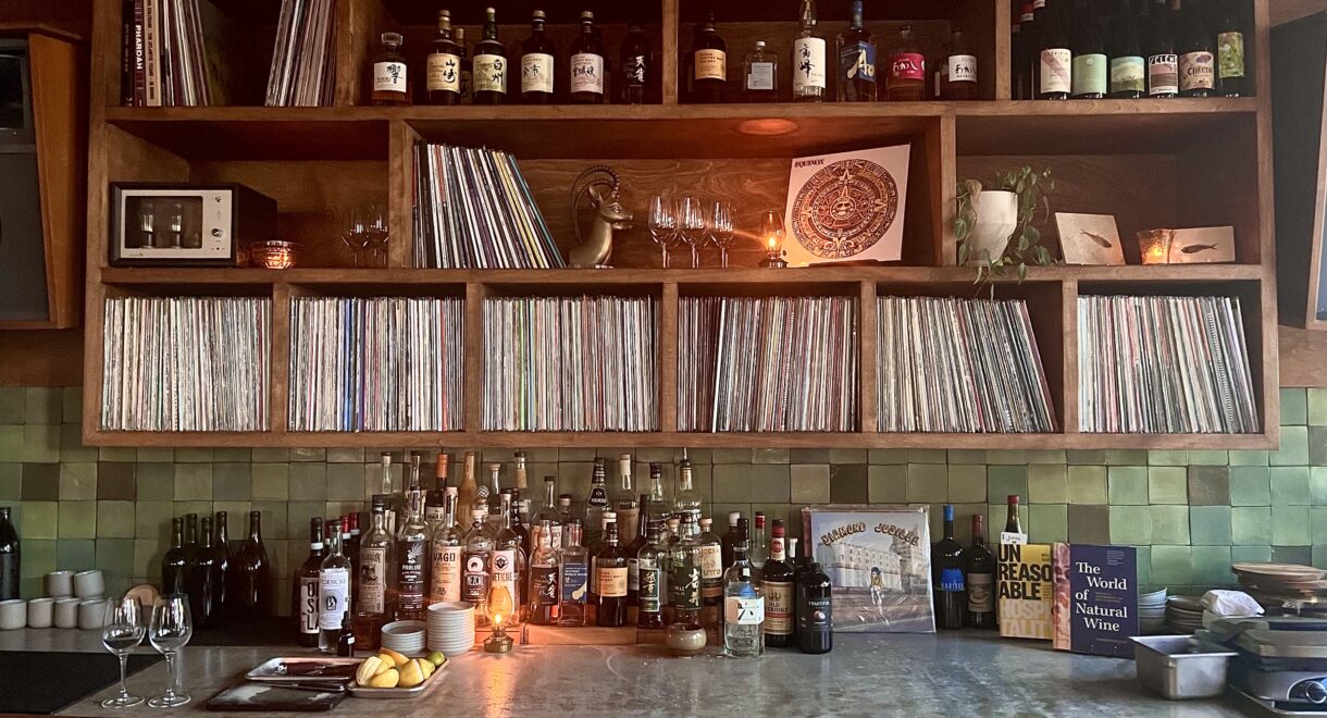 Close-up of XO’s back bar, where shelves are lined with vinyl records, bottles of spirits, and books. The middle section features rows of LPs organized tightly together, flanked by candles, wine glasses, and small decorative objects. Bottles of whiskey, mezcal, and other liquors sit on the counter below alongside lemons, glassware, and cocktail tools. On the right, two books are displayed upright—Unreasonable Hospitality and The World of Natural Wine—next to a record sleeve propped against the green-tiled backsplash. Warm lighting creates a cozy, intimate atmosphere.