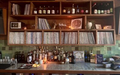 Close-up of XO’s back bar, where shelves are lined with vinyl records, bottles of spirits, and books. The middle section features rows of LPs organized tightly together, flanked by candles, wine glasses, and small decorative objects. Bottles of whiskey, mezcal, and other liquors sit on the counter below alongside lemons, glassware, and cocktail tools. On the right, two books are displayed upright—Unreasonable Hospitality and The World of Natural Wine—next to a record sleeve propped against the green-tiled backsplash. Warm lighting creates a cozy, intimate atmosphere.