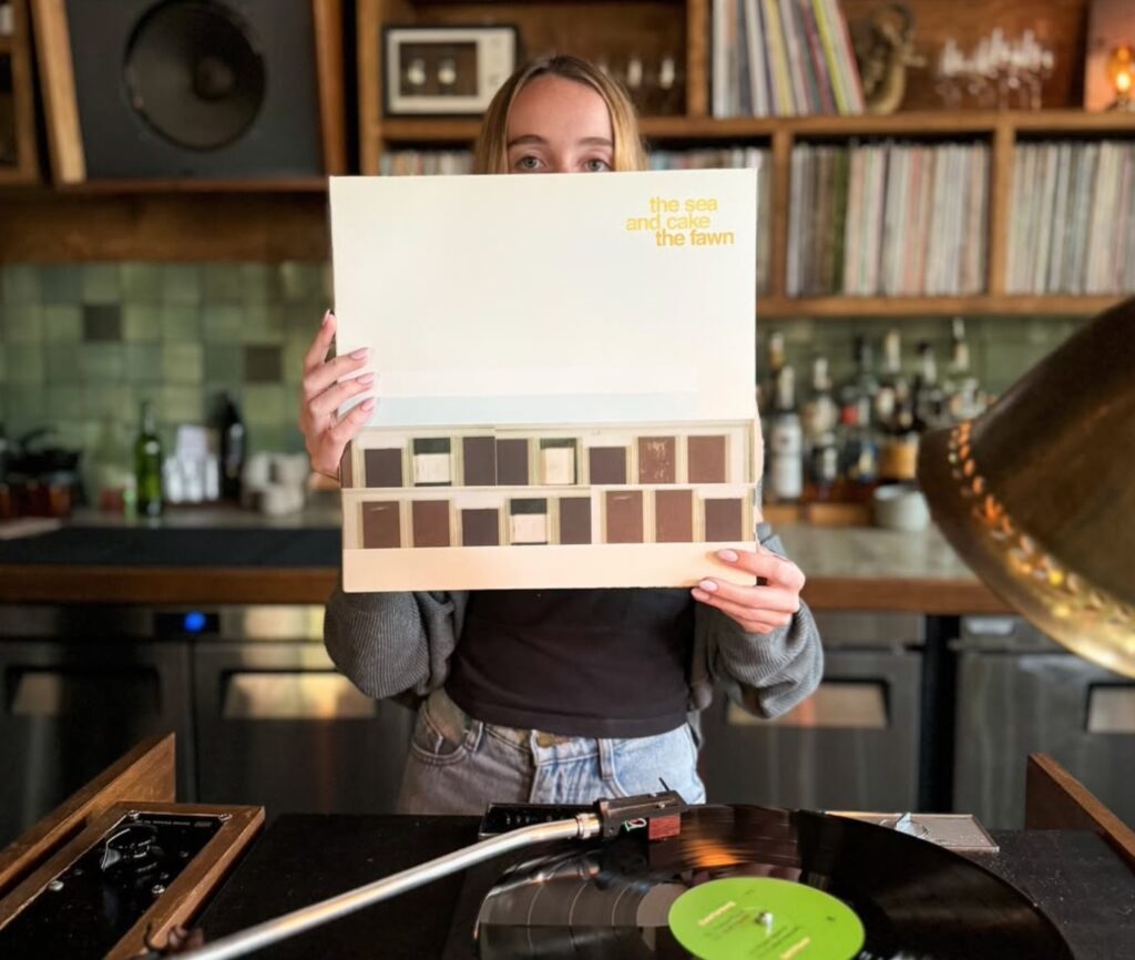 A person stands behind the bar at XO holding up a vinyl copy of The Fawn by The Sea and Cake, partially obscuring their face. The cream-colored album cover features minimalist block graphics and pale lettering. In the foreground, a turntable with a record spinning on the platter is visible, its green label catching the light. Behind the person, shelves are lined with rows of LPs, bottles of spirits, and small candles, set against a backdrop of green tile. The photo captures the listening bar’s blend of music and atmosphere.