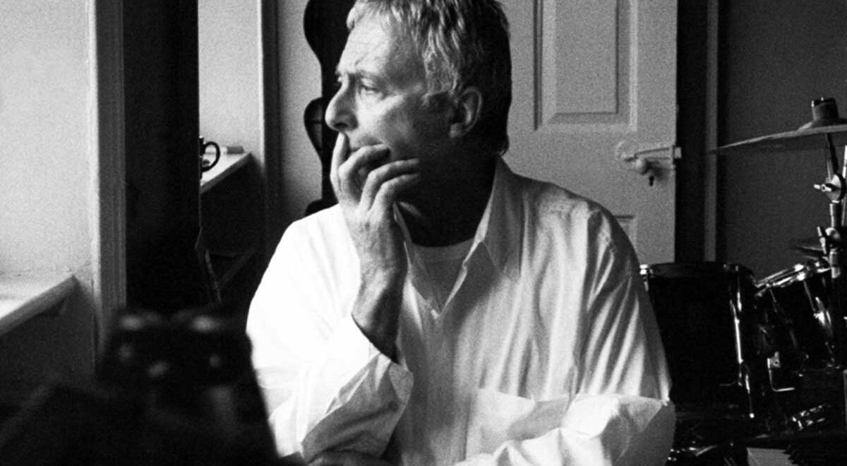 Black-and-white portrait of composer Harold Budd sitting by a window, gazing thoughtfully into the light with his hand resting against his chin. He wears a white button-down shirt, and a drum kit and keyboard are partially visible behind him, suggesting a quiet studio setting.