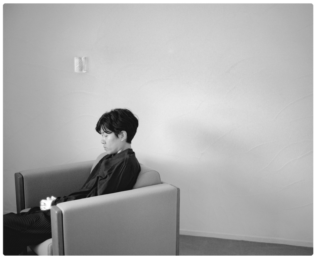 Black-and-white photograph of a person seated alone in a modern armchair, viewed in profile against a plain textured wall. They wear dark clothing and look down thoughtfully, hands clasped, the soft light emphasizing the quiet, contemplative atmosphere of the room.