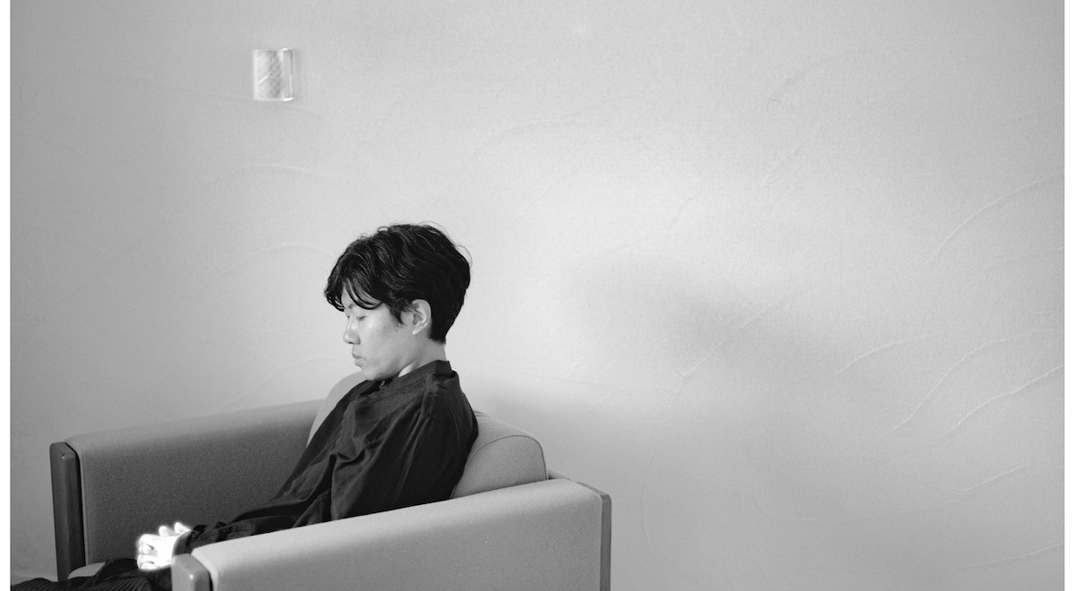 Black-and-white photograph of a person seated alone in a modern armchair, viewed in profile against a plain textured wall. They wear dark clothing and look down thoughtfully, hands clasped, the soft light emphasizing the quiet, contemplative atmosphere of the room.
