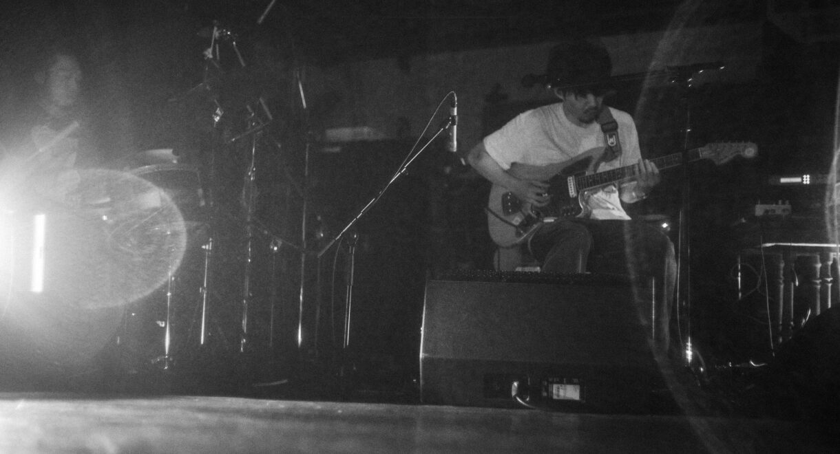 A calm, moody black-and-white photograph of musician Takuro Okada seated in low light, looking down as he plays an electric guitar. His face is partly in shadow, and the soft grain of the image emphasizes the quiet, intimate atmosphere of the performance.
