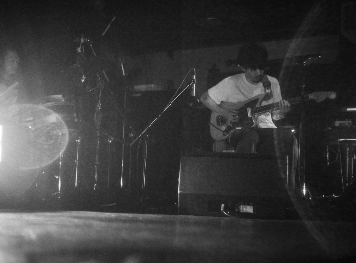 A calm, moody black-and-white photograph of musician Takuro Okada seated in low light, looking down as he plays an electric guitar. His face is partly in shadow, and the soft grain of the image emphasizes the quiet, intimate atmosphere of the performance.