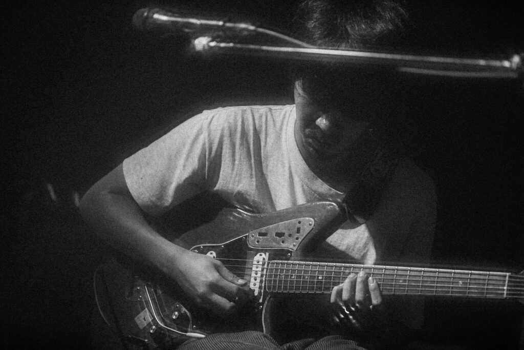 A calm, moody black-and-white photograph of musician Takuro Okada seated in low light, looking down as he plays an electric guitar. His face is partly in shadow, and the soft grain of the image emphasizes the quiet, intimate atmosphere of the performance.