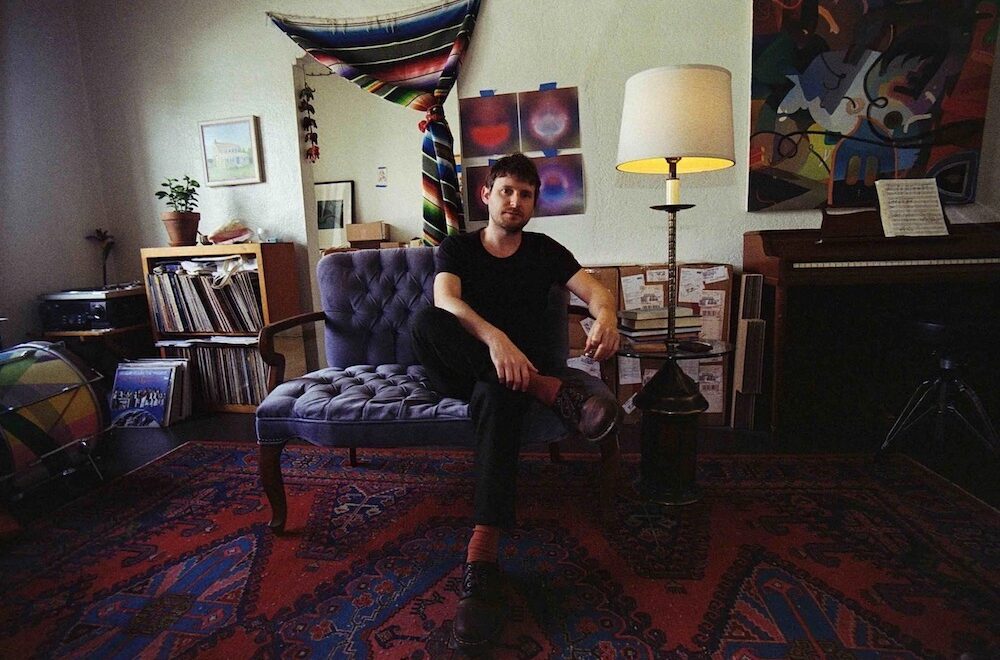 Brendan Eder sits on a tufted purple loveseat in a warmly lit living room studio, surrounded by a piano, stacked records, books, and abstract artwork. He faces the camera with a relaxed posture, one leg crossed, set against patterned rugs and a tall floor lamp. Photo by Laura Moreau.