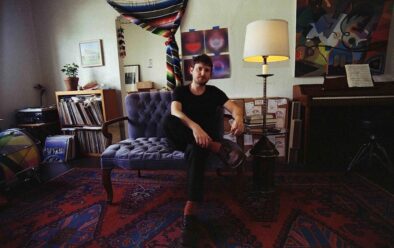 Brendan Eder sits on a tufted purple loveseat in a warmly lit living room studio, surrounded by a piano, stacked records, books, and abstract artwork. He faces the camera with a relaxed posture, one leg crossed, set against patterned rugs and a tall floor lamp. Photo by Laura Moreau.
