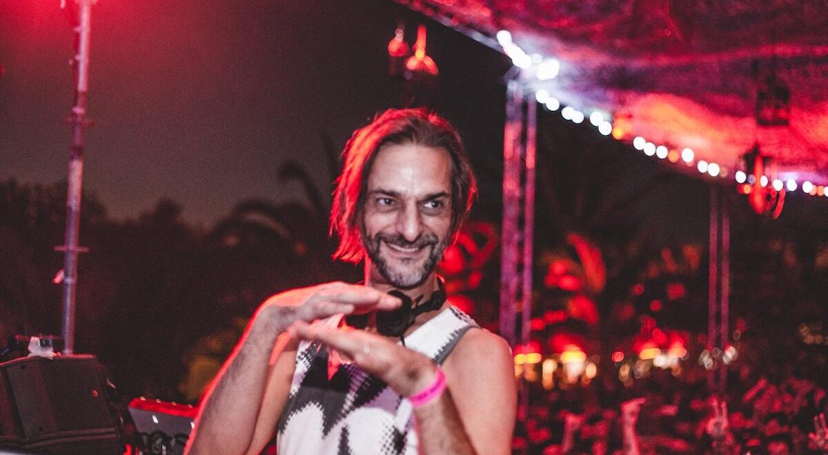 A smiling DJ Ricardo Villalobos stands behind turntables under red stage lighting, wearing a sleeveless patterned top and headphones around his neck, with one hand lifted mid-gesture as a crowd and palm trees blur behind him in a lively nighttime outdoor setting.