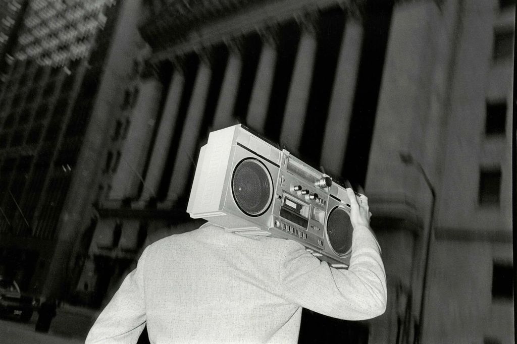 Black-and-white photo of a person seen from behind walking through a city at night, carrying a large vintage boombox on their shoulder. Tall buildings rise on either side, their vertical lines slightly blurred, while the figure’s light-colored jacket and the angular shape of the stereo stand out against the darker urban backdrop.
