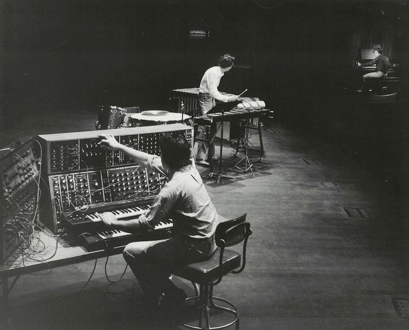 Black-and-white photo of three musicians performing onstage: one seated at a large modular synthesizer in the foreground, another standing at a vibraphone or percussion setup in the center, and a third at a piano in the background. The arrangement emphasizes the scale of the analog equipment and the spatial relationship between performers in a dim, rehearsal-like environment.