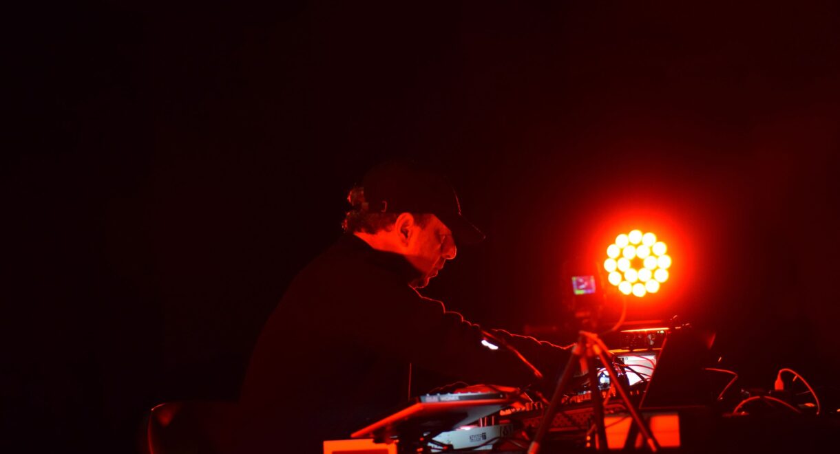 Musician Michel Banabila performs at an electronic setup on a dark stage, illuminated by intense red lighting. Wearing a black cap and jacket, he leans over a table of synthesizers, mixers and cables while adjusting controls. A bright circular LED light glows behind him, casting a red halo and leaving most of the surrounding space in deep shadow.