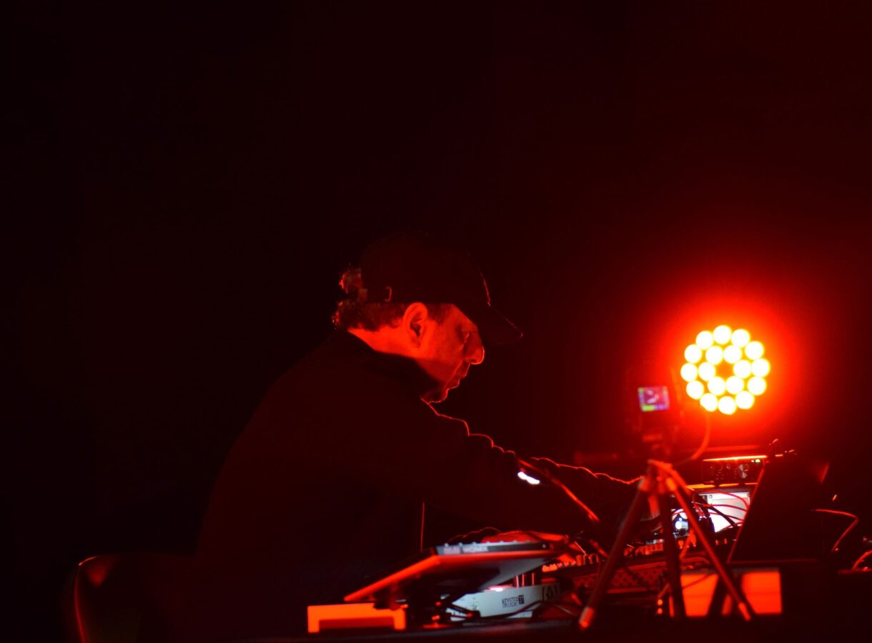 Musician Michel Banabila performs at an electronic setup on a dark stage, illuminated by intense red lighting. Wearing a black cap and jacket, he leans over a table of synthesizers, mixers and cables while adjusting controls. A bright circular LED light glows behind him, casting a red halo and leaving most of the surrounding space in deep shadow.