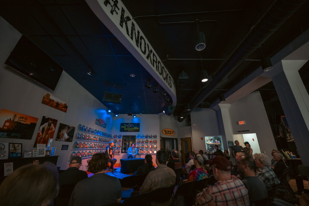 A seated audience fills a small, dimly lit gallery space during a live Essential Tremors podcast recording at Big Ears. Two speakers sit onstage under blue lighting, facing each other in conversation at a low table, while rows of attendees watch attentively. A large curved banner reading “Knoxville Rocks” hangs overhead, and the walls are lined with circular displays and local music signage, reinforcing the intimate, community-driven setting.