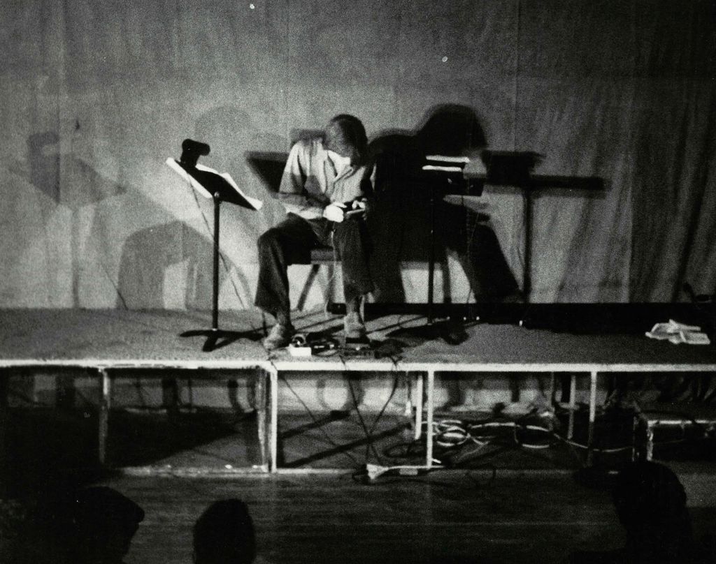 Black-and-white photo of a solo performer seated on a small stage, leaning forward over a compact setup of electronic devices and cables. A music stand sits to the left, while stark lighting casts oversized shadows of the performer and equipment across a plain backdrop, giving the scene a quiet, experimental feel.