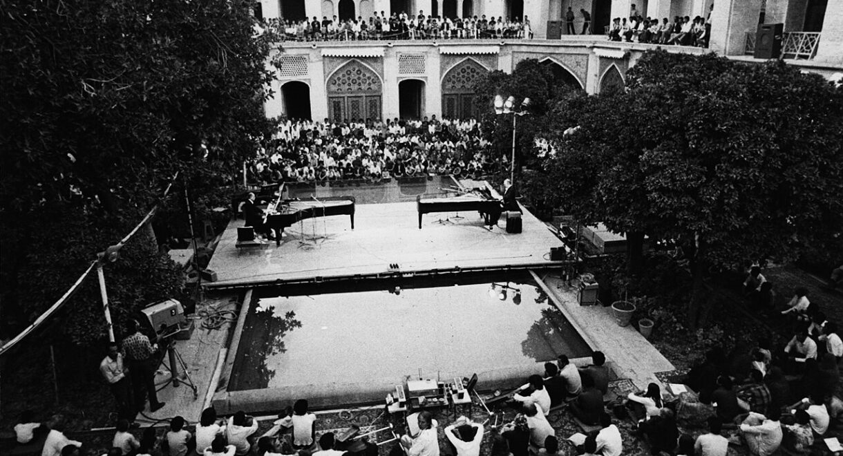 Black-and-white photograph of a performance of Mantra by pianists Aloys and Alfons Kontarsky at Seraye Moshir during the Shiraz Arts Festival, September 2, 1972. Two grand pianos face each other on a raised stage set over a rectangular reflecting pool in a historic courtyard. A large audience fills the surrounding arcaded balconies and sits closely packed on the ground level, watching the evening concert beneath open sky.