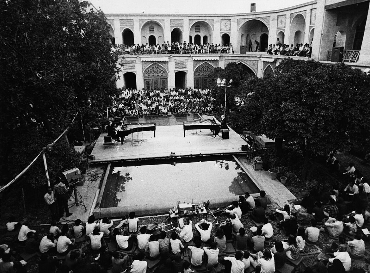 Black-and-white photograph of a performance of Mantra by pianists Aloys and Alfons Kontarsky at Seraye Moshir during the Shiraz Arts Festival, September 2, 1972. Two grand pianos face each other on a raised stage set over a rectangular reflecting pool in a historic courtyard. A large audience fills the surrounding arcaded balconies and sits closely packed on the ground level, watching the evening concert beneath open sky.