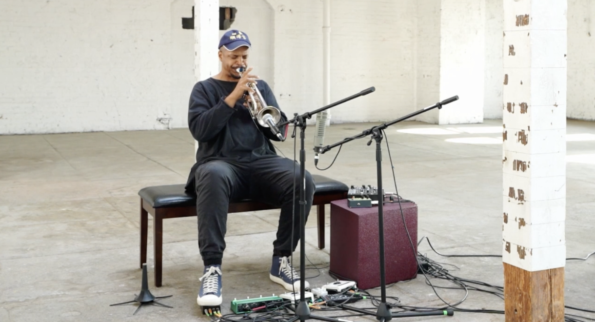Chris Williams sits on a bench in a sparse, sunlit industrial space, playing trumpet into a pair of microphones, with cables and effects pedals spread across the concrete floor at his feet.