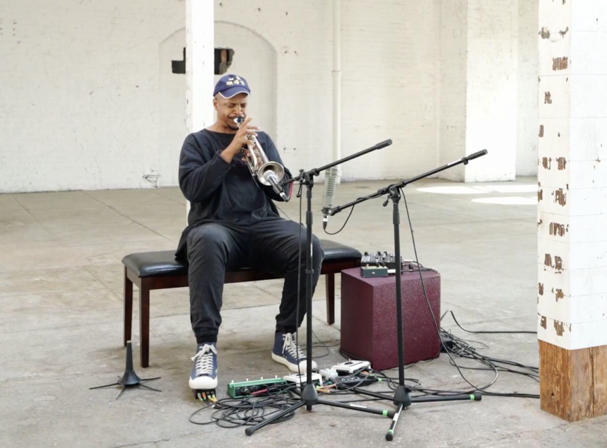 Chris Williams sits on a bench in a sparse, sunlit industrial space, playing trumpet into a pair of microphones, with cables and effects pedals spread across the concrete floor at his feet.