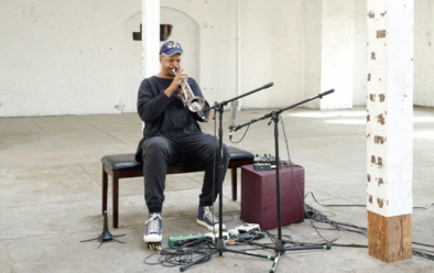 Chris Williams sits on a bench in a sparse, sunlit industrial space, playing trumpet into a pair of microphones, with cables and effects pedals spread across the concrete floor at his feet.