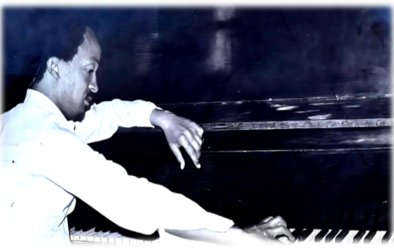 Black-and-white portrait of Wesenyeleh Mebreku seated at a keyboard, his body angled in profile as he leans forward with one hand extended across the keys. His posture is focused and intent, suggesting mid-performance or rehearsal, while the softly lit background fades into darkness, drawing attention to his hands and the instrument.