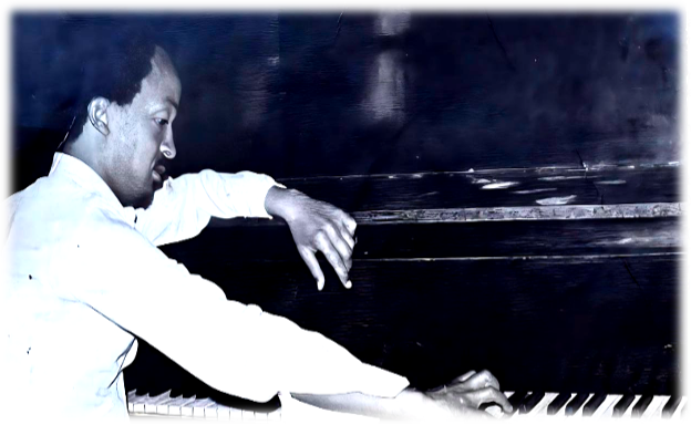 Black-and-white portrait of Wesenyeleh Mebreku seated at a keyboard, his body angled in profile as he leans forward with one hand extended across the keys. His posture is focused and intent, suggesting mid-performance or rehearsal, while the softly lit background fades into darkness, drawing attention to his hands and the instrument.