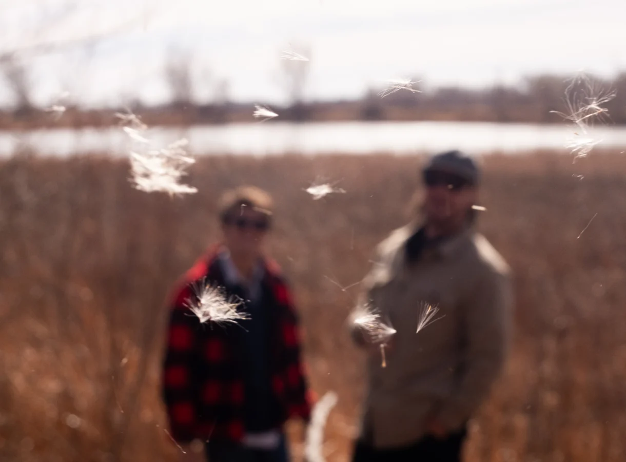 Soft-focus image of two figures standing in a dry field near water, their forms blurred in the background while delicate white seeds drift sharply in the foreground, suspended midair in warm, late-day light.